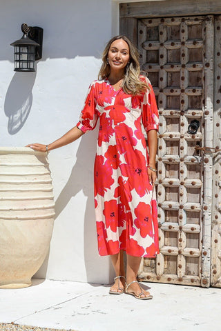 Woman wearing a red and white floral midi dress and sandals standing by a rustic wooden door.