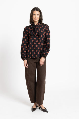 Woman wearing a black polka dot long sleeve blouse with a bow neck and brown trousers.