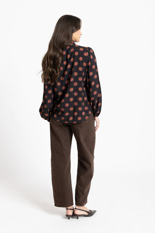Woman wearing a black long-sleeve blouse with large brown polka dots and brown wide-leg pants.