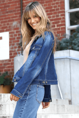 Smiling woman wearing a blue denim jacket and matching denim jeans standing outdoors.