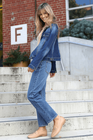 Smiling woman wearing a denim jacket, blue jeans, and tan loafers on concrete stairs.
