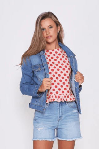 Woman wearing a red polka dot top, blue denim jacket, and light wash denim shorts.