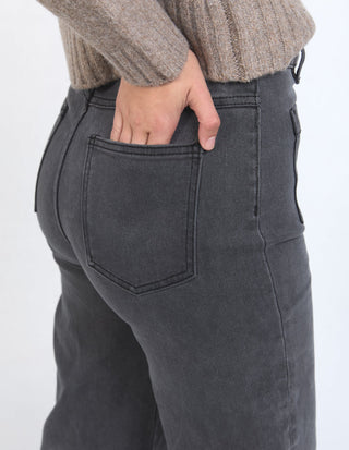 Close-up of a person wearing charcoal grey denim pants with a hand in the back pocket.