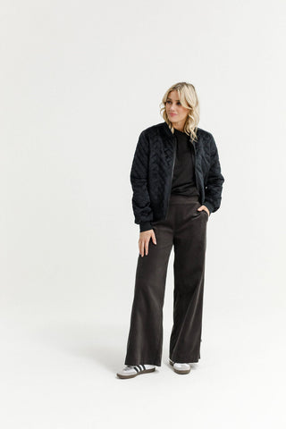 Woman wearing a black quilted bomber jacket, black top, and dark wide-leg trousers.