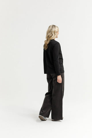 Woman wearing a black long-sleeve top and wide-leg black trousers walking against a white background.