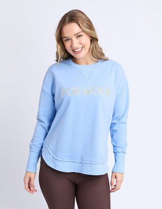 Woman smiling in a light blue Foxwood sweatshirt and brown pants.