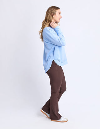 Woman wearing a light blue sweatshirt and brown flared pants standing in profile.