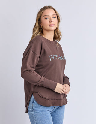 Woman wearing a brown Foxwood long sleeve sweatshirt and blue jeans.