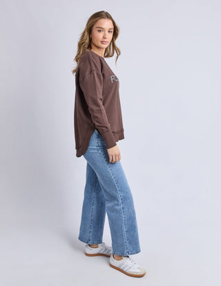 Woman wearing a brown Foxwood sweatshirt and blue wide-leg denim jeans with white sneakers.