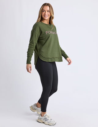 Woman wearing an olive green Foxwood sweatshirt, black leggings, and white sneakers.