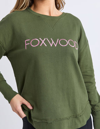 Woman wearing an olive green Foxwood sweatshirt with pink embroidered lettering.