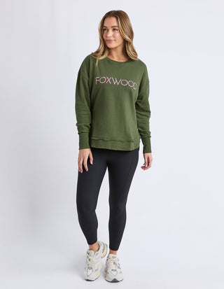 Woman wearing an olive green Foxwood sweatshirt, black leggings, and white sneakers.
