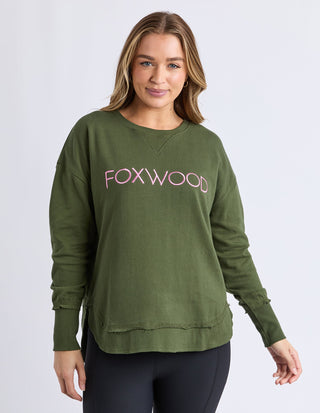Woman wearing an olive green Foxwood sweatshirt with pink lettering and black leggings.