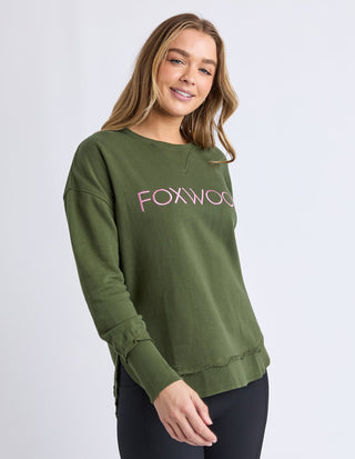 Woman wearing an olive green Foxwood sweatshirt with pink lettering and black leggings.