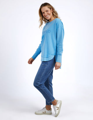 Woman wearing a blue Foxwood sweatshirt and denim jeans with white sneakers.
