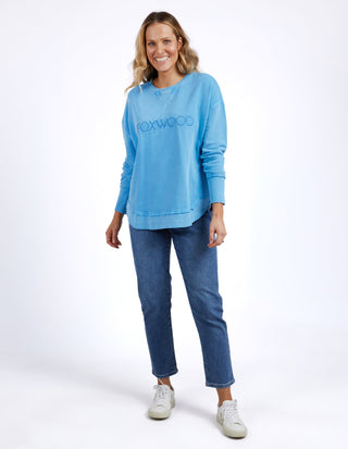 Woman smiling in a blue Foxwood sweatshirt and blue denim jeans with white sneakers.