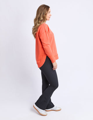 Woman wearing a bright orange sweatshirt and charcoal grey flare pants with white sneakers.