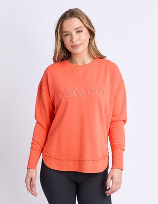 Woman wearing an orange Foxwood sweatshirt with a raw edge hem and black leggings.