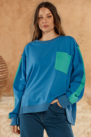 Woman wearing an oversized blue sweatshirt with green accent pocket and sleeve details.