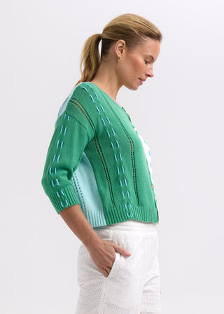 Woman wearing a green and blue knit cardigan with white eyelet trousers.