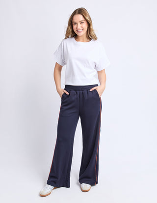 Smiling woman wearing a white t-shirt and navy blue wide-leg lounge pants with orange side stripes.